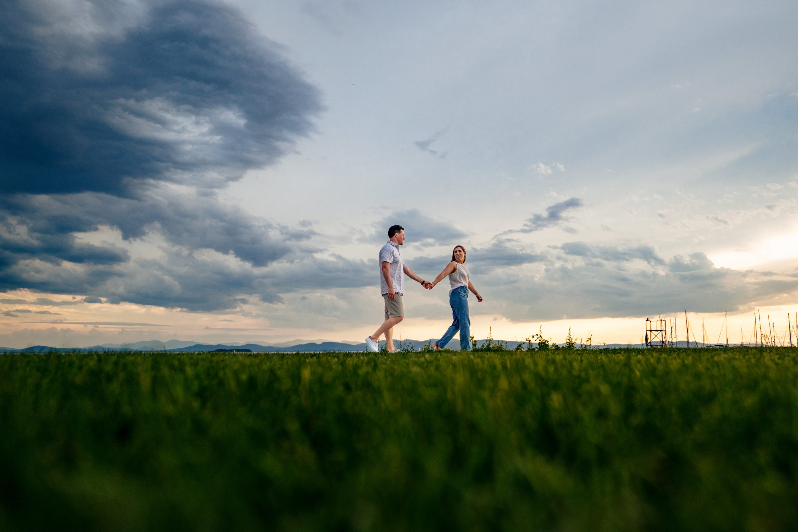 sunset engagement photography at burlington vermont waterfront. Couple walking through the grass with a summer sunset in the background