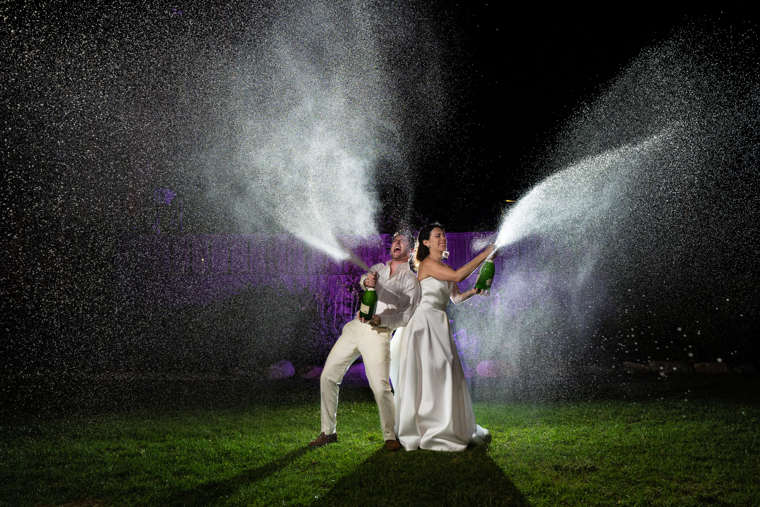 Bride and groom celebrate their wedding night by spraying champagne under the night sky, with dramatic lighting and joyful expressions.